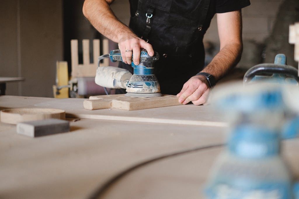 sanding with an orbital sander. Unrecognizable male woodworker wearing uniform and polishing wooden board with random orbital sander at big table in professional studio