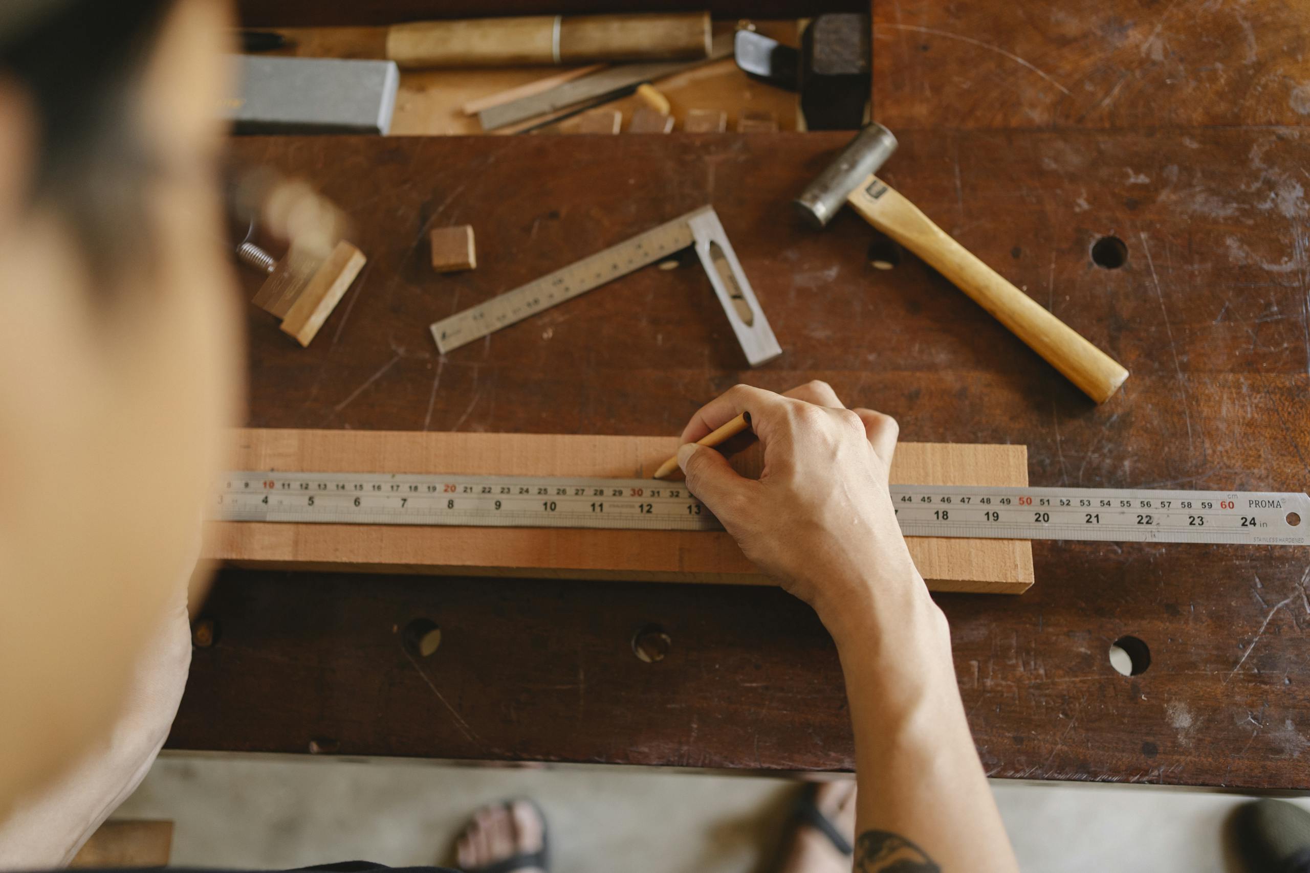 Top view of crop anonymous male measuring timber plank with ruler and marking with pencil while working on table with tools